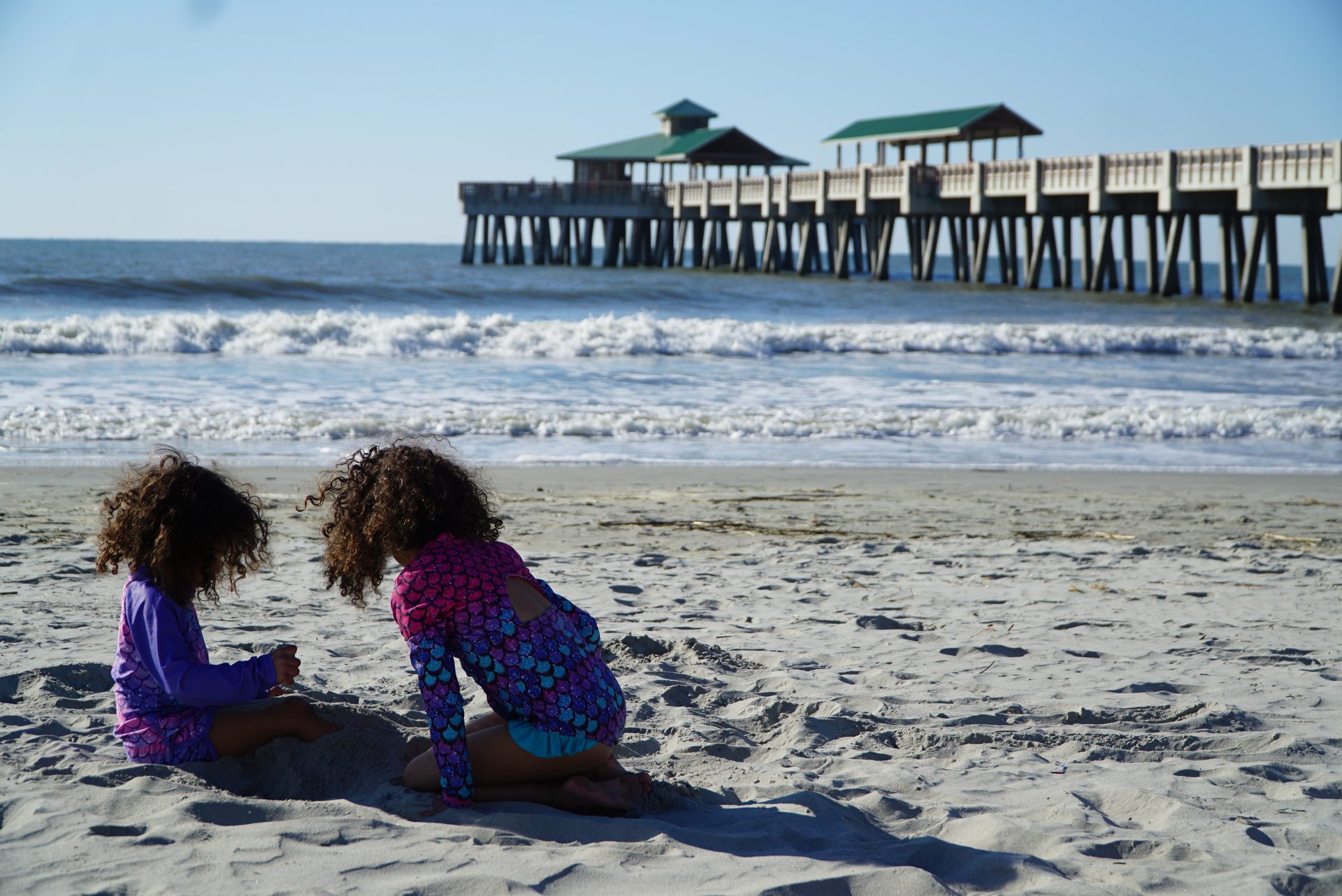 Two children sitting from behind, looking out at the waves and the Folly Beach pier