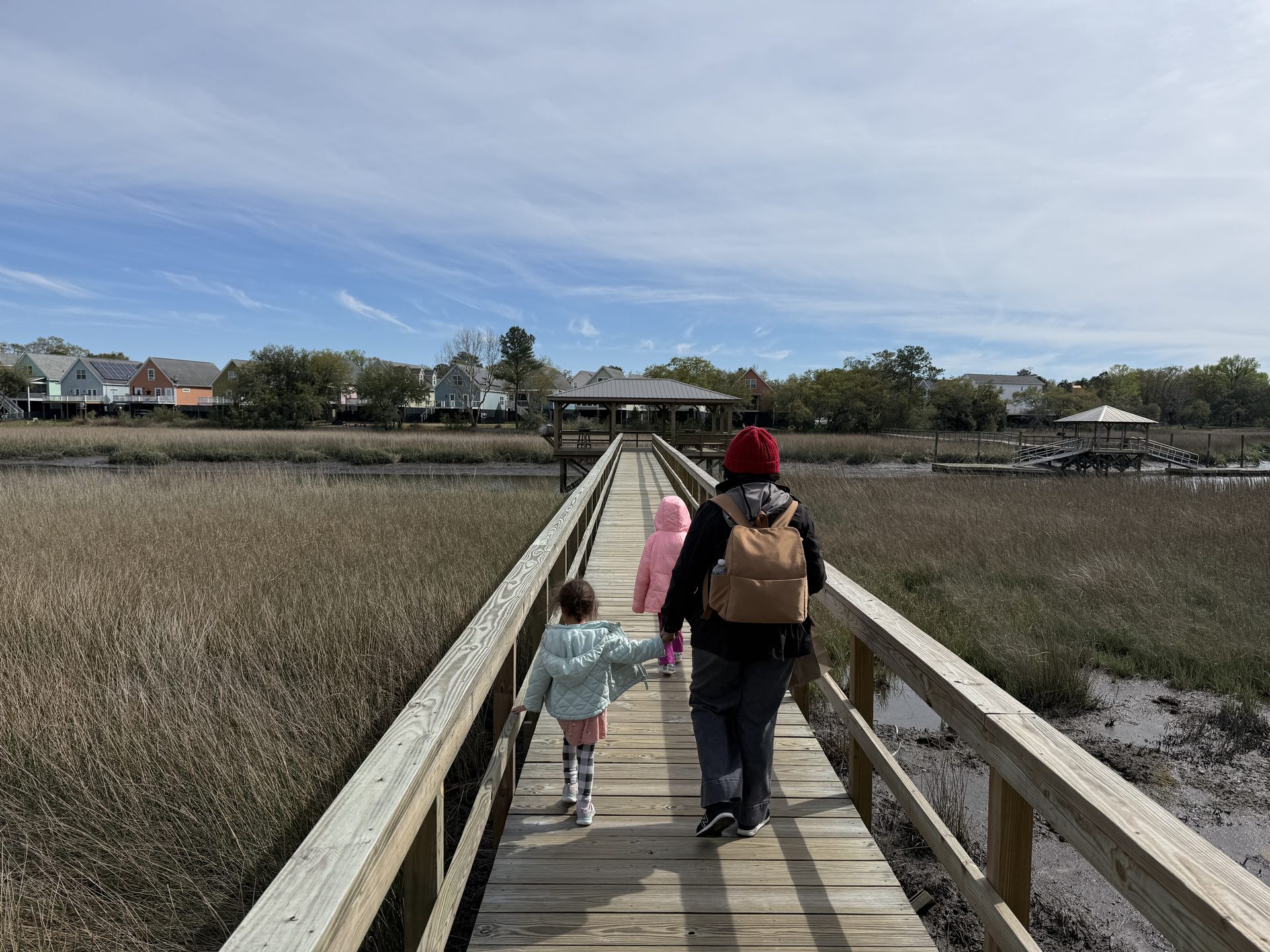 Adult and child walking hand-in-hand down a long wooden boardwalk over a tidal marsh at Holy City Brewing